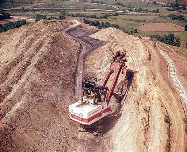 The Gem of Egypt working its way through the Egypt Valley strip mine of Ohio in the late 1960s early 1970s. The  giant machine is removing the earth over the coal seam on the right, and then, swinging its boom and loaded shovel  left, depositing the “overburden” on the spoil banks. A tiny vehicle mid-photo appears to be riding on a road that is  the unearthed coal seam. At right, atop the hillside being mined, is a line of powdered-white blasting holes where dynamite will be used to loosen the “overburden” the giant shovel will continue to remove. 
