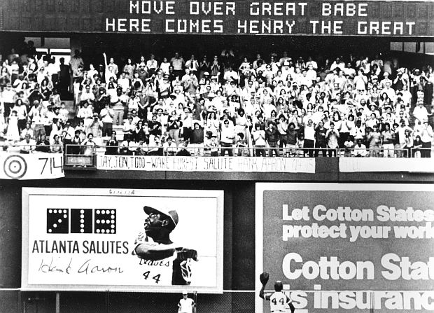 Photo of Henry Aaron – No. 44, small figure, bottom center, at his position in the outfield during game – waving to, and acknowledging fans’ ovation for him at Atlanta Braves stadium on the last game of the season, even though he had 713, but unable to tie or surpass the Babe Ruth record during his at bats in that last game of 1973.