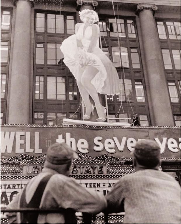Huge cut-out figure of Marilyn Monroe in her famous "subway grate" pose is set into position above a Lowes Theater in New York City to promote the 1955 film, The Seven Year Itch, as two workmen observe the scene..