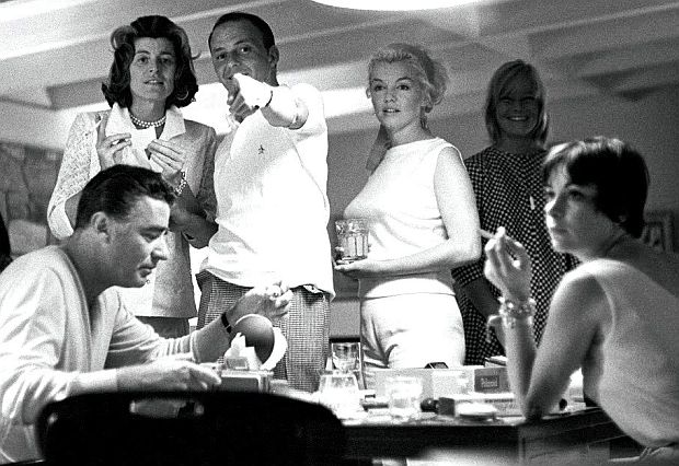 Photo of the same group as above, now showing Patricia Kennedy Lawford standing at left with Sinatra pointing, Peter Lawford at left now seated, and Shirley MacLaine at right, turning toward camera.