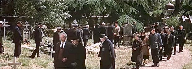 At the cemetery, a steady line of mourners & admirers attend the burial service for Zhivago, surprising half-brother Yevgraf (standing at center red collar), not realizing the popular appeal of his brother’s poetry. 